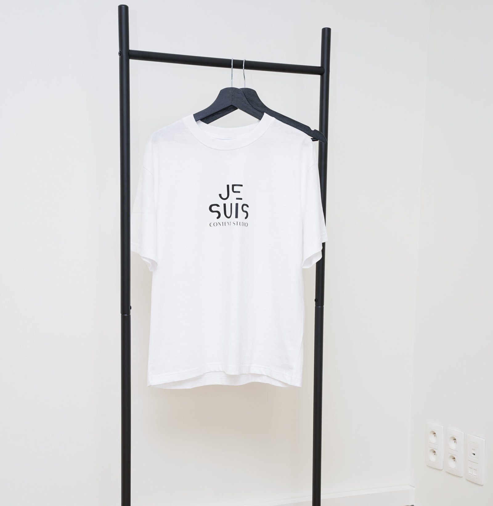 White Je Suis Content Studio branded t-shirt hanging on black clothing rack in Antwerp studio